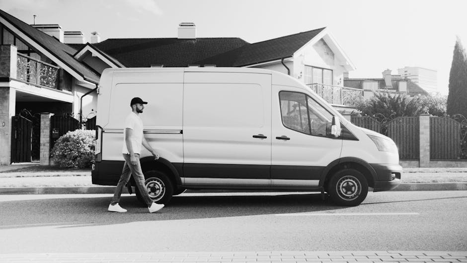 A man wearing casual clothing, including a cap and white sneakers, walking alongside a white cargo van parked on a residential street. The van is positioned parallel to the pavement, with its sliding