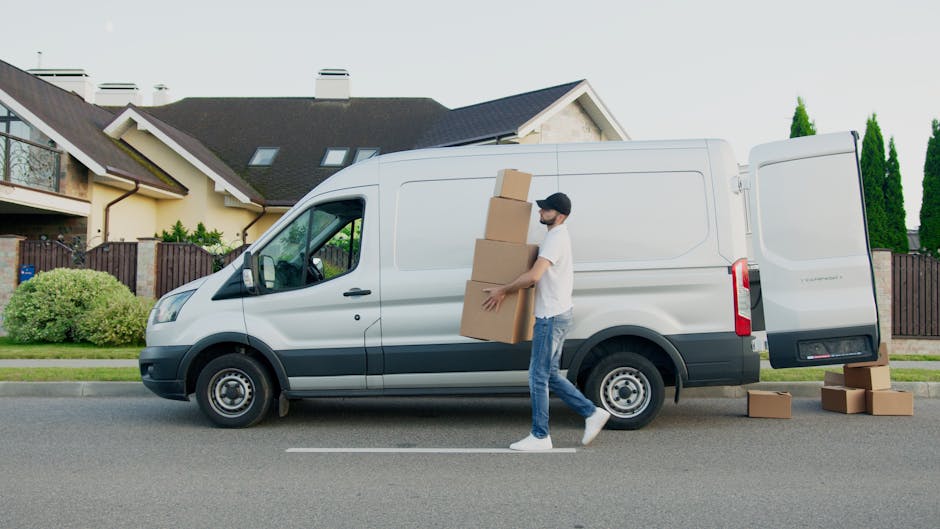 A man wearing casual clothing, including a cap and white sneakers, walking alongside a white cargo van parked on a residential street. The van is positioned parallel to the pavement, with its sliding