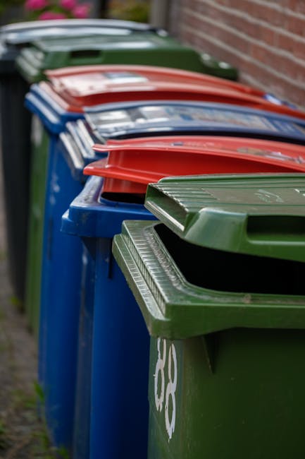 A row of three large wheelie bins placed on a paved sidewalk in front of a residential property, with two black bins, one blue bin, and one green bin, each labeled with recycling or waste management symbols. The bins are positioned near a black metal lamppost and are partially blocked by fallen autumn leaves scattered on the ground. Behind the bins, there is a tall, dense green hedge, alongside a wooden fence and the exterior of a modern white house with black-framed windows. The scene is outdoors on a quiet residential street with trees showing early signs of autumn foliage, and a car visible parked in the distance along the curb. The image depicts a typical setting for household waste disposal and is relevant to the logistics of house clearance and recycling during home relocation or removals, as managed by companies like Man with Van Pimlico.