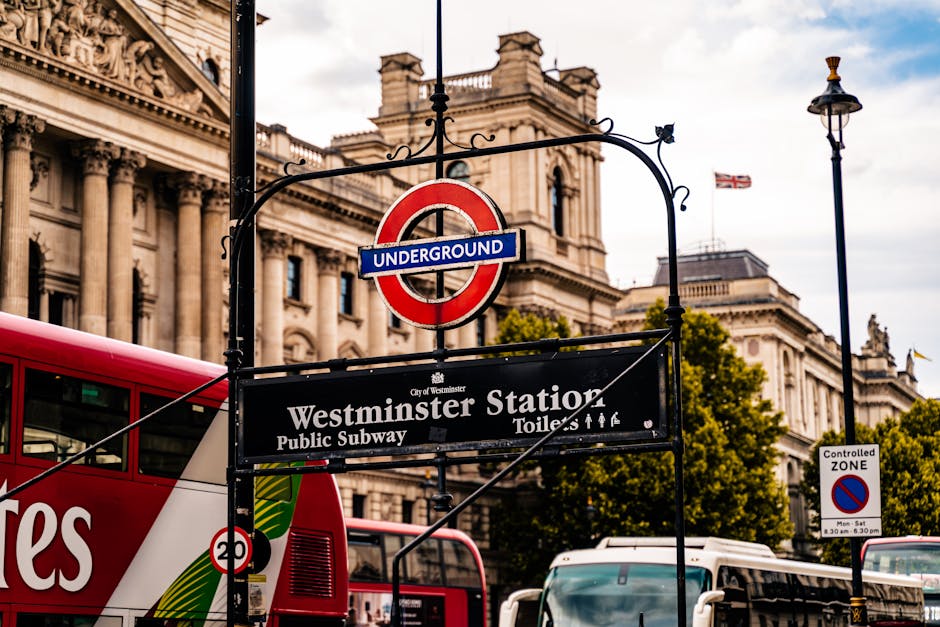 A street scene in Pimlico showing a London Underground sign for Westminster Station hanging above a black signboard that indicates public subway, toilets, and staff, with an image of a red double-decker bus partly visible below. In the background, there is a historic building with ornate architectural details, large windows, and decorative columns, along with a flag flying atop the structure. On the pavement and near the building, trees with green foliage are visible, and a streetlamp stands to the right. The scene is captured during daylight with partly cloudy skies, reflecting typical urban surroundings in Westminster. The context relates to house removals and the logistics of moving within or around Westminster, with natural lighting and an emphasis on street infrastructure supporting relocation activities, as seen through the signage and urban environment.