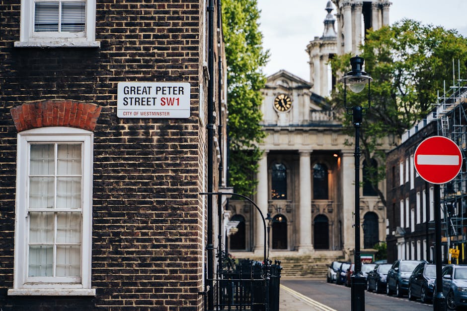 A street scene on Great Peter Street in Westminster with a close-up of a dark brick building featuring a white-framed window partly covered by a red brick arch, a street sign indicating the location as Westminster, SW1 postal district, and a street lamp. Behind this, there is a view of a grand classical-style building with large columns and a clock, surrounded by trees with green leaves. Parked cars are visible along the curb on the right side of the street, and a red and white 'No Entry' traffic sign is positioned on the corner. The setting suggests an urban area suitable for house removals or furniture transport, with the scene capturing the environment where professional movers like Man with Van Pimlico often operate during the loading, packing, and home relocation processes in a historic city district.