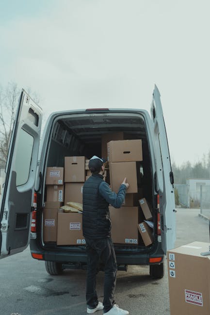 Two individuals, a man and a woman, are engaged in a house relocation process involving furniture transport and packing. They are positioned at the open rear of a white moving van, which is parked out