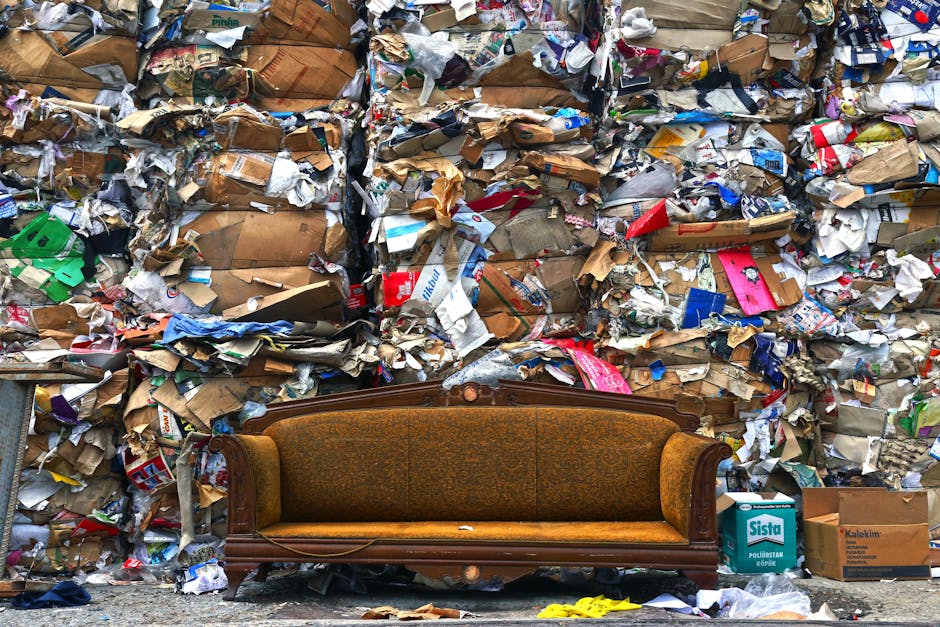 A vintage upholstered sofa with a wooden frame and floral fabric in a mustard and brown color scheme is positioned on a pavement in front of a large, densely packed pile of mixed waste materials, including flattened cardboard boxes, crumpled paper, plastic packaging, and cardboard cartons. The waste appears to be outside a property, possibly after a house clearance or disposal of bulky waste. The background consists of the cluttered waste pile, which extends above the sofa, indicating a significant quantity of discarded packaging and waste. The scene is illuminated by natural daylight, highlighting the textures of the furniture and the rough, chaotic arrangement of the accumulated waste. The image captures the process of clearing and removal of household waste, relevant to professional removals and waste disposal services such as those offered by Man with Van Pimlico, who assist in house removals and bulky waste clearance during home relocation or decluttering projects.