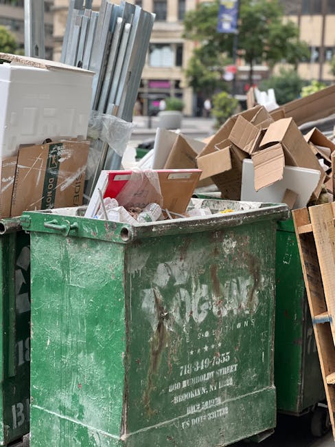 A large green waste disposal bin, positioned outdoors on a street, filled with cardboard boxes, plastic packaging, and other household waste materials. The contents include flattened cardboard shipping cartons, some with visible tape and printing, and smaller white plastic packaging. The bin is surrounded by other waste containers, with some boxes stacked nearby, and an urban environment with buildings, trees, and pedestrians in the background. This scene depicts the process of clearing bulky waste after a home removal or decluttering, as part of a house clearance or moving logistics handled by Man with Van Pimlico, who specializes in removals and transport services, including waste disposal and furniture transport.