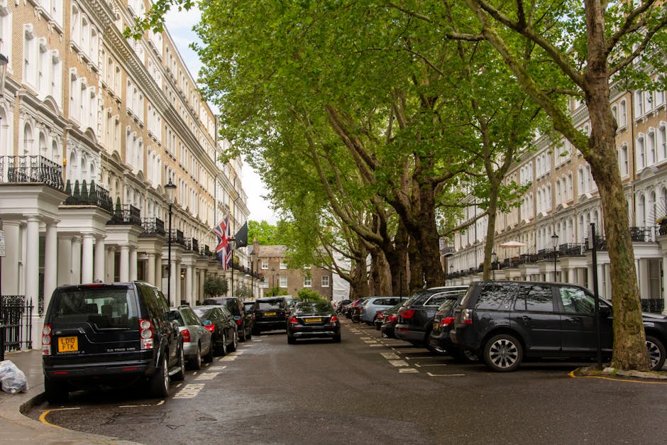 A street scene in SW1V with a row of white Victorian-style terraced houses featuring black wrought-iron balconies and flagpoles displaying the Union Jack and a black flag. The street is lined with large, leafy green trees that extend their branches over the pavement and parked vehicles, creating a canopy. Numerous cars, including vans and sedans, are parked along on both sides of the narrow street, some partially on the pavement. The street surface appears wet, suggesting recent rain. In the distance, a black vehicle is seen moving away from the camera, near the entrance of the loading bay of a property used for home relocation and furniture transport. Some packaged boxes and moving materials are visible near the curb, indicating packing activities associated with house removals. This scene exemplifies an organized, residential moving environment, with a focus on parking and loading logistics, consistent with the services offered by Man with Van Pimlico.