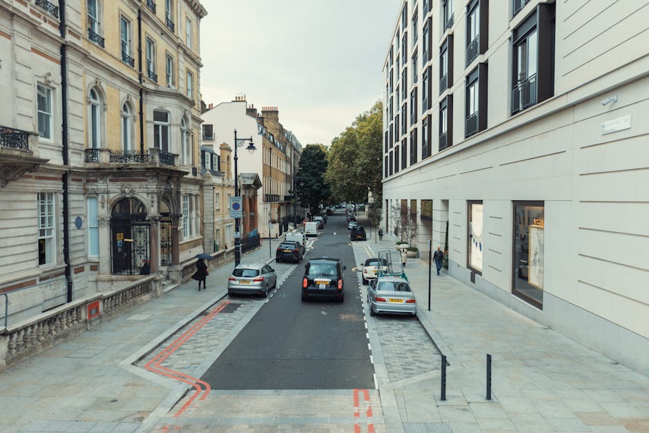 A photo taken from an elevated perspective showing a city street with parked cars lining both sides of the road, amidst historic and modern buildings in Pimlico, SW1V. The street features a designated loading bay area with red markings on the pavement, indicating parking restrictions or loading zones. The loading bay is currently empty, with surrounding clear pavement for vehicle access. Pedestrians are walking along the sidewalks, some approaching the buildings. The scene includes a large, modern building on the right with multiple windows and a neutral façade, while on the left, there are older, ornate buildings with decorative balconies and arched windows. In the background, a few trees and additional parked vehicles are visible, with natural daylight illuminating the scene, suggesting daytime. This setting illustrates urban logistics relevant to house and furniture removals, where careful loading and unloading take place in a well-organized environment, characteristic of professional removal services like Man with Van Pimlico.