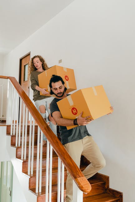 Two individuals are ascending a narrow staircase inside a residential property, each carrying a large cardboard box with packing tape, which are marked with a red 'H' label. The man in the foreground, dressed in a grey t-shirt and beige trousers, is holding a box close to his chest while carefully navigating the steps. Behind him, a woman with curly hair, wearing a brown jacket and ripped jeans, is also carrying a box and following him up the staircase. The staircase has wooden steps and a polished wooden handrail on the left side, with white walls framing the scene. This image captures the process of home relocation, where careful handling of packed boxes is essential for safe furniture transport and packing during the moving or relocation services provided by Man with Van Pimlico. The lighting is natural, highlighting the interior of a well-maintained flat, emphasizing the logistical steps involved in moving small household items along narrow interior staircases.
