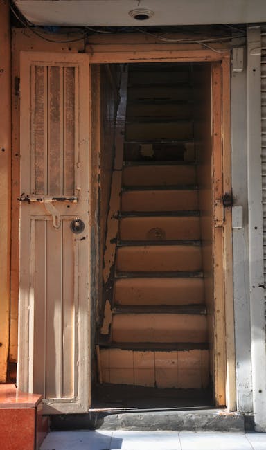 A narrow staircase inside a property with wooden steps and an unfinished, rough wooden frame around the doorway. The stairs are situated behind a doorway that appears to lead to the staircase, with visible gaps and unpainted surfaces. On the left side of the doorway, there is a small wooden door panel with a bolt latch and a circular keyhole, attached to a wooden frame. The surrounding area shows exposed construction elements, with some metal supports and a partially visible metallic shutter or panel on the right side. The scene is lit by natural or ambient light, and the environment suggests ongoing renovation or house relocation work, involving careful handling of furniture and boxes, supported by Man with Van Pimlico’s removal services.