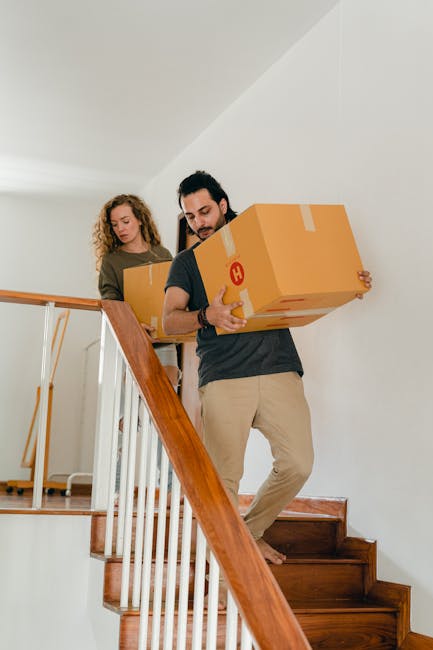 Two individuals are ascending a narrow staircase inside a residential property, each carrying a large cardboard box with packing tape, which are marked with a red 'H' label. The man in the foreground, dressed in a grey t-shirt and beige trousers, is holding a box close to his chest while carefully navigating the steps. Behind him, a woman with curly hair, wearing a brown jacket and ripped jeans, is also carrying a box and following him up the staircase. The staircase has wooden steps and a polished wooden handrail on the left side, with white walls framing the scene. This image captures the process of home relocation, where careful handling of packed boxes is essential for safe furniture transport and packing during the moving or relocation services provided by Man with Van Pimlico. The lighting is natural, highlighting the interior of a well-maintained flat, emphasizing the logistical steps involved in moving small household items along narrow interior staircases.