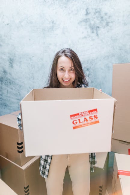 Inside a residential property, a woman with long brown hair is smiling and holding an open cardboard moving box labeled 'GLASS' in red text, indicating fragile contents. She is dressed in casual clothing, with a plaid shirt and light-colored pants. The background features a textured blue wall, and several other cardboard boxes of various sizes, some sealed with red or black tape, are arranged on the floor around her. One box appears to contain packing materials, possibly paper or bubble wrap. The scene captures a packing or moving process, with the woman preparing for home relocation. The environment suggests a well-lit indoor space, with ample natural or artificial light illuminating the area, and the focus is on the careful handling of packing items as part of furniture transport or packing and moving activities undertaken by Man with Van Pimlico for swift, same-day removals in SW1.