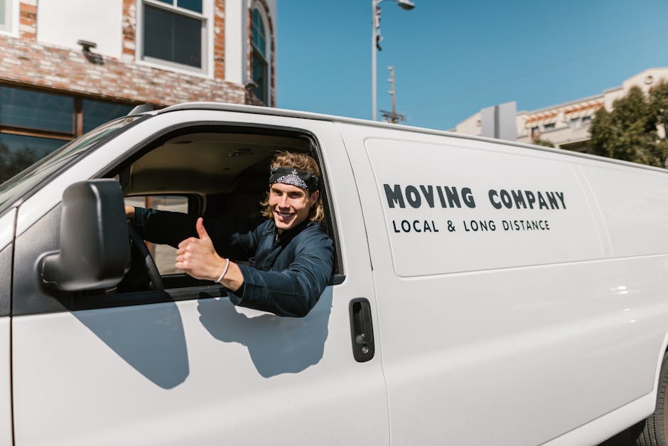 A smiling young man with long hair, wearing a black headband and dark clothing, is sitting in the driver’s seat of a white moving van, visible through the open window. The van features the company name 'Moving Company' and the words 'Local & Long Distance' printed on the side. Inside the vehicle, various packing materials, such as cardboard boxes and protective padding, are visible near the cabin area, indicating a home relocation and furniture transport process. The van is parked on a street with a clear blue sky overhead, and nearby buildings with brick and glass facades are partially visible in the background. This scene depicts the loading and transportation phase of a professional removals service, consistent with house moving procedures carried out by [COMPANY_NAME], with an emphasis on logistics, packing, and safe vehicle loading.
